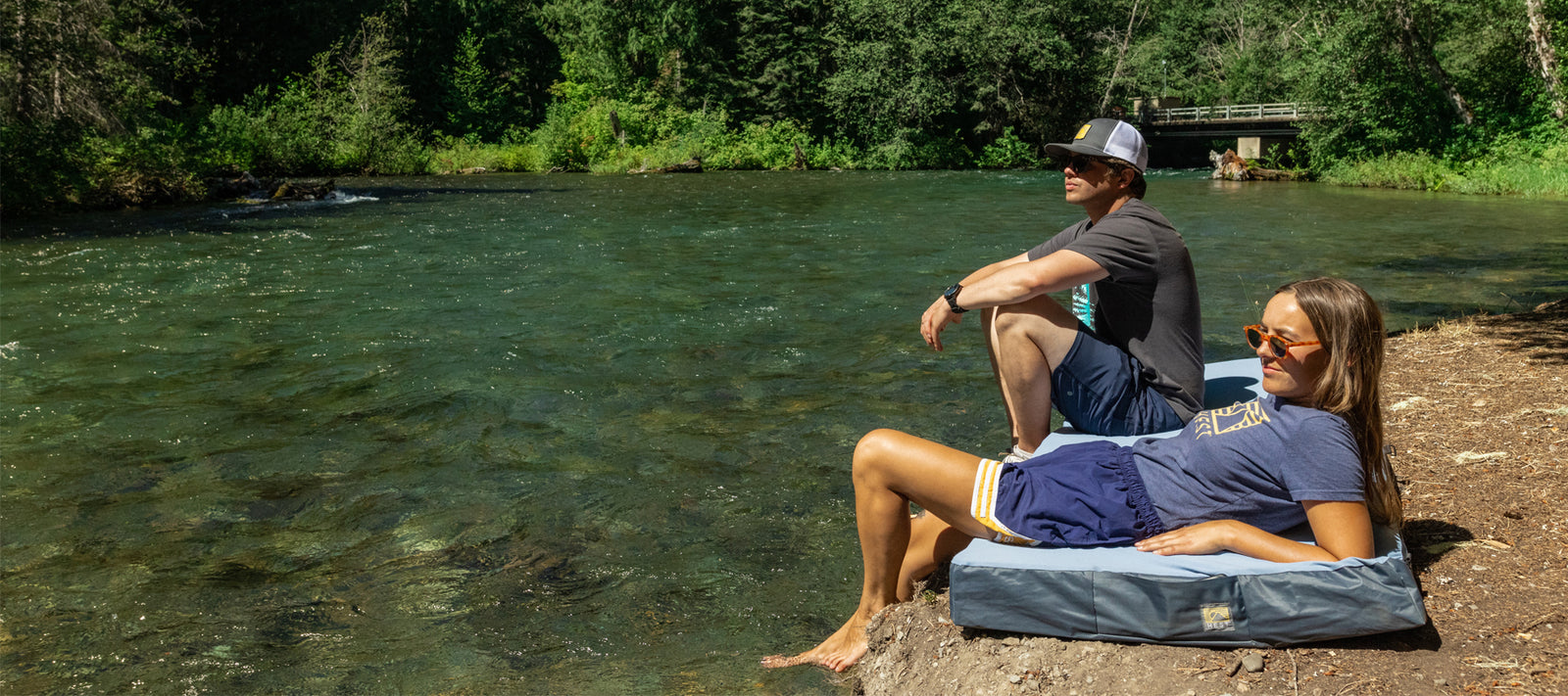 HEST's top tips to staying cool this summer and beat the heat. Image features two people sitting on a comfortable camping mattress next to a river. Their feet are dipped in the river. It is very sunny and bright and hot in the photo.